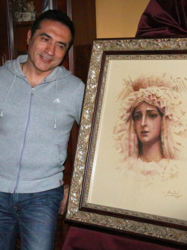 Un hombre sonríe mientras sostiene un cuadro de una mujer con vestimenta tradicional. La imagen muestra detalles de la decoración interior, incluyendo una estantería con libros y un tapiz.