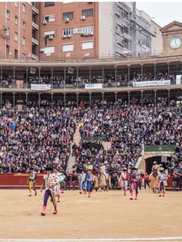 Un torneo de tauromaquia en una plaza de toros, con toreros y vaqueros en el centro del ruedo. La multitud de espectadores está sentada en las gradas, observando el evento. En la parte superior del ruedo se puede ver una torre con una gran reloj. La imagen está tomada en un día soleado, con el cielo claro y las sombras de los espectadores.