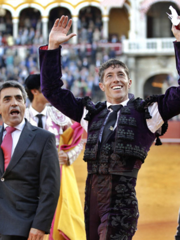 Un torero en traje de lujo celebra con un puño levantado, mientras otro torero y un hombre en traje de gala observan. El escenario es una plaza de toros con asistentes en los arcos y un fondo de espectadores.