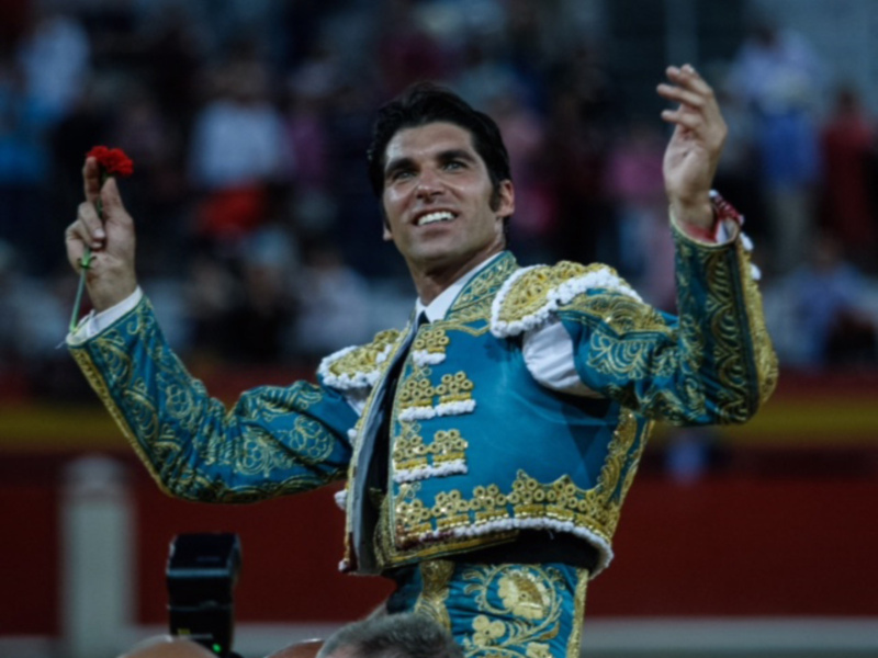 Un torero en traje tradicional de color azul y dorado, con un rostro sonriente y una mano levantada sosteniendo una flor roja. El fondo muestra un público en la arena de toros, con algunos fotógrafos capturando el momento.