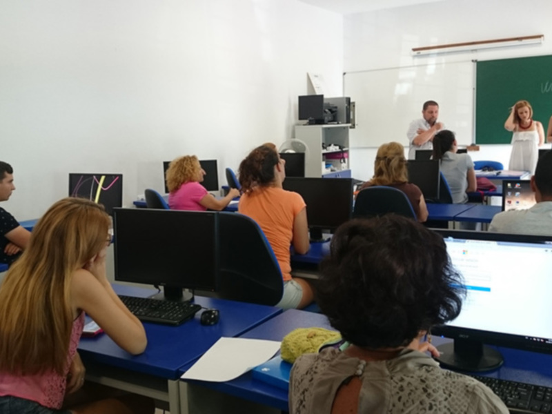 Clase de informática en un aula con estudiantes usando computadoras. Un instructor está explicando algo, mientras otros estudiantes trabajan en sus propios computadores. La sala tiene una pizarra y un proyector.