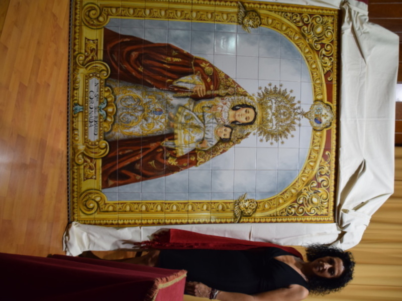 Una imagen de una mujer sosteniendo un tapiz con una representación religiosa en el centro, rodeada por detalles dorados y rojos.