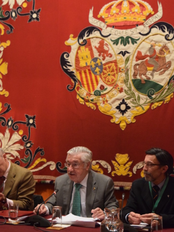 Imagen de una reunión formal con tres hombres sentados en una mesa frente a un escudo heráldico español. El ambiente es formal y el fondo muestra una bandera roja con un escudo heráldico.