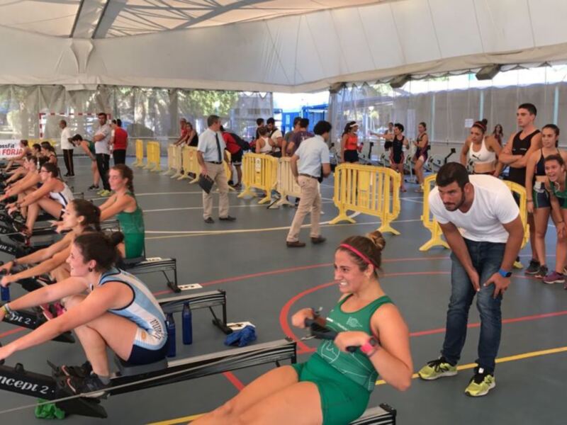 Una imagen de un grupo de atletas en una sesión de entrenamiento indoor, sentados en bicicletas de remo. El ambiente es dinámico con personas en el fondo, algunos con equipos deportivos y otros observando. La iluminación es natural gracias a la luz del día que entra por las ventanas, y el suelo es de color azul con líneas rojas que delimitan los espacios. La estructura superior es blanca, probablemente una cubierta para proteger a los atletas del clima.