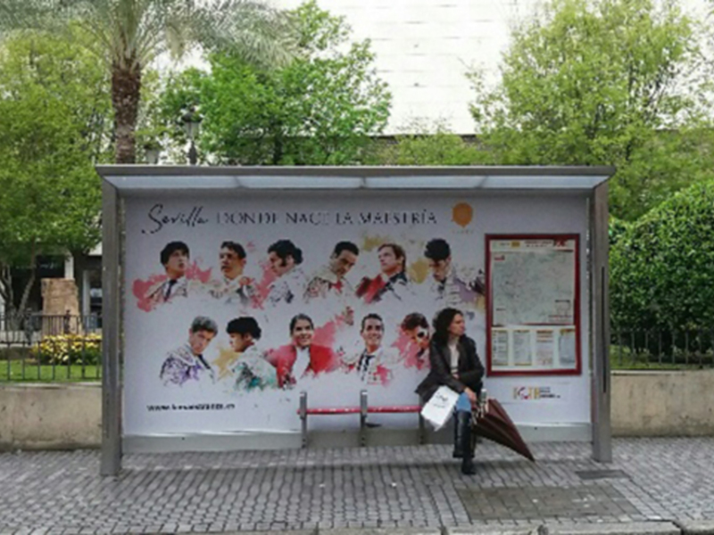 En un parque, una persona se sienta en un banco de la estación de autobuses. Al lado, hay una publicidad con imágenes de personas y texto en español sobre Sevilla. La estación está rodeada por árboles y una cerca de metal.
