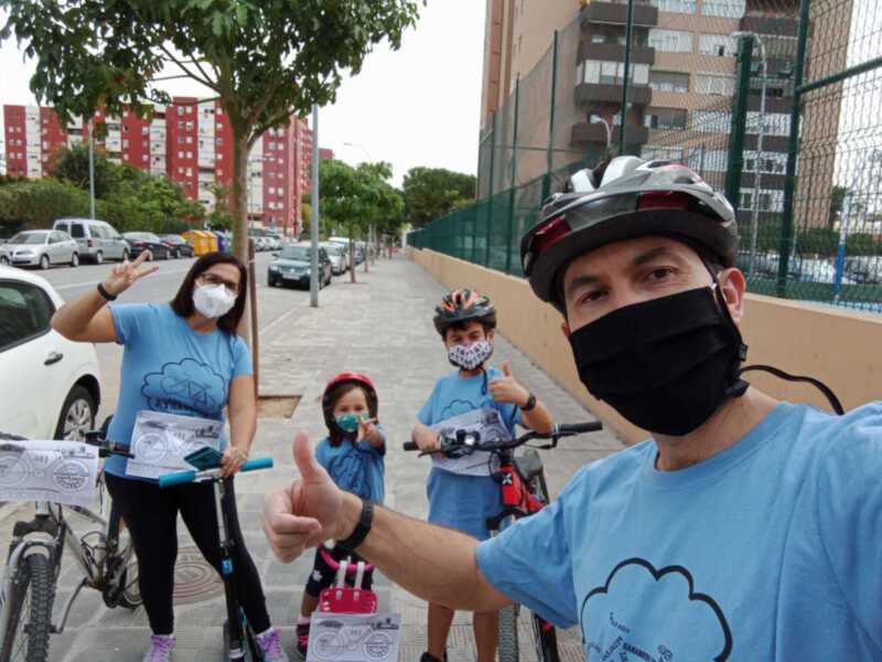 Una familia en bicicleta y patineta, todos con cascos y mascarillas. El padre da un gesto de aprobación mientras la madre y los hijos sonríen. Fondo urbano con edificios y árboles.