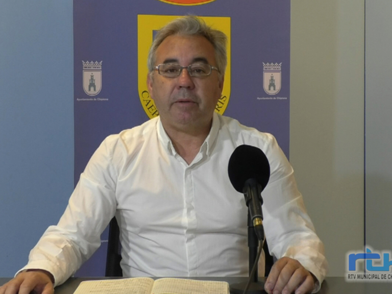 El hombre está sentado en una mesa con un libro abierto frente a él, hablando por un micrófono. La pared detrás de él tiene una bandera española y el escudo del Ayuntamiento de Chipiona.
