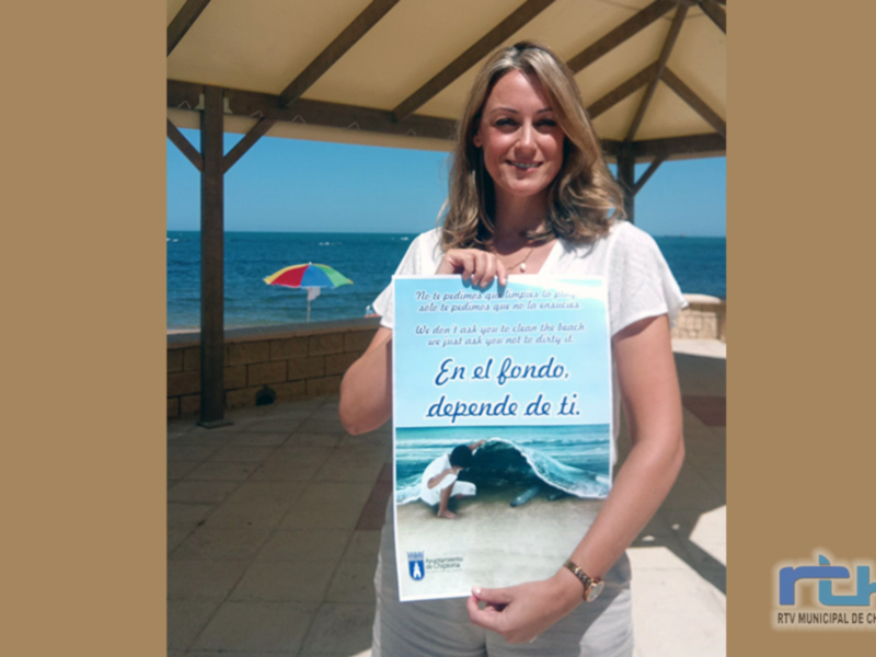 Una mujer con cabello largo y rubio sostiene un folleto sobre el fondo del mar. La imagen se encuentra en una estructura con techo de madera, frente a un océano tranquilo con una playa y una sombrilla multicolor en el fondo.