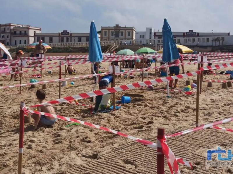 Una playa con juegos infantiles y personas jugando. Frontera de seguridad roja y blanca en el fondo, edificios al fondo.
