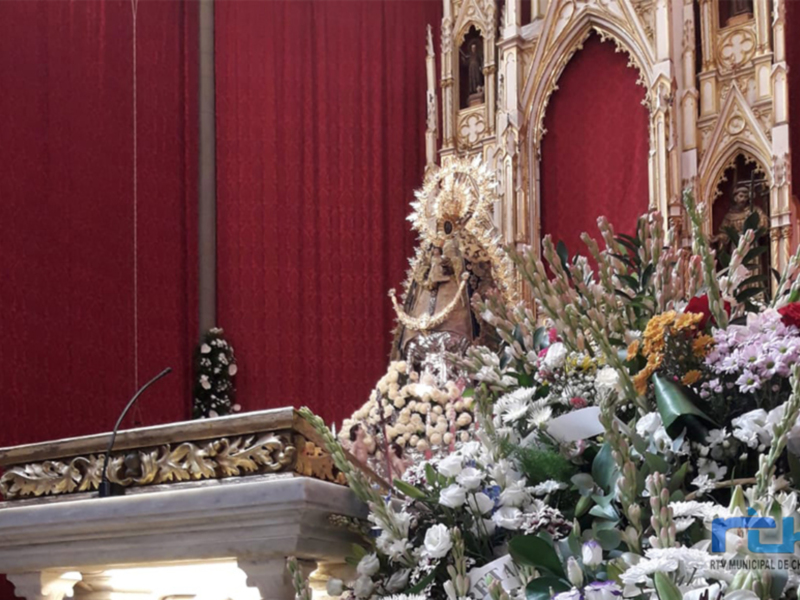 Una escultura religiosa en un altar rodeada de flores y adornos dorados. El fondo es una pared roja con detalles dorados, creando un ambiente de solemnidad y reverencia.