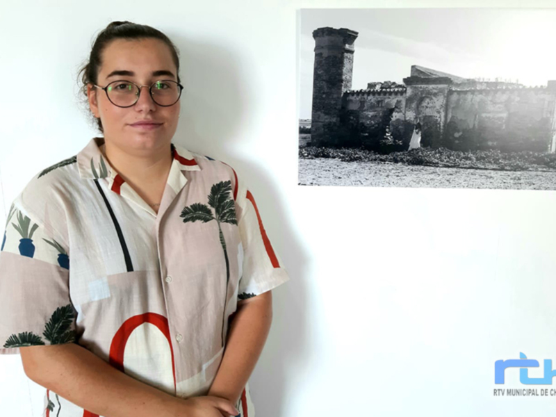 A una persona con cabello largo y gafas, vestida con un camiseta de manga corta con diseño tropical. A su lado, hay una foto antigua en blanco y negro de un edificio histórico con torres. La imagen tiene el logo de RTV Municipal de Chipiona en la esquina inferior derecha.