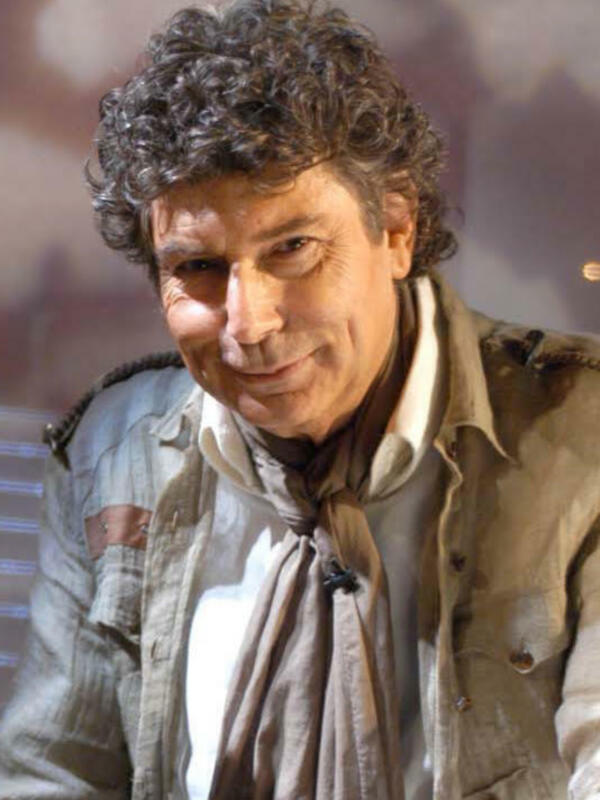 Un hombre con cabello rizado y ojos grandes, sonriendo. Está vestido con una chaqueta beige y un jersey blanco. La imagen parece ser de una entrevista o programa de televisión.