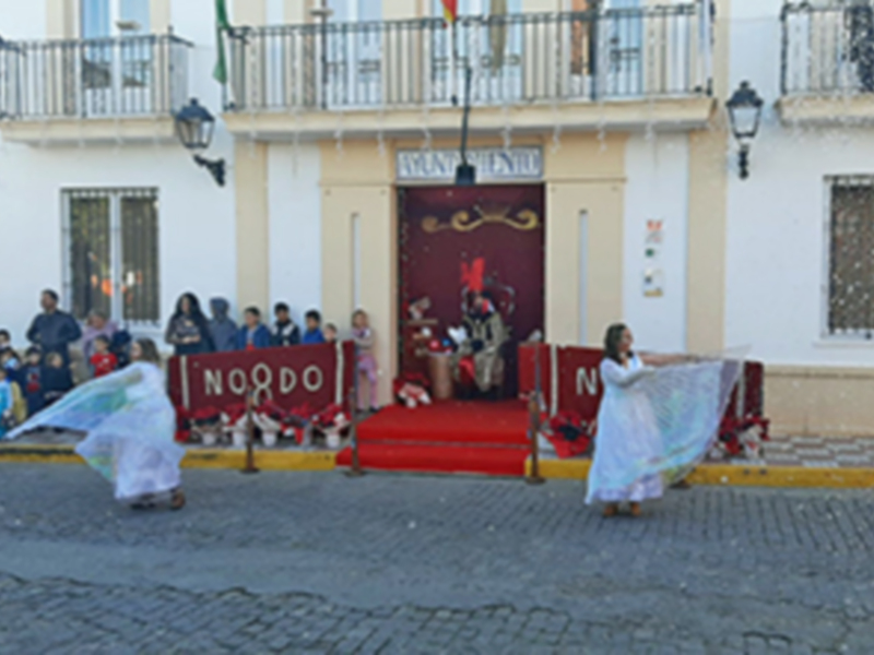 Personas desfilando con mantones en una calle de España, frente a un edificio blanco y rojo.