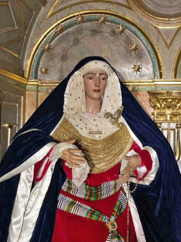 Una estatua de la Virgen María vestida con un manto azul y una saya roja, adornada con cintas de colores. La estatua está colocada en un entorno decorativo con molduras doradas y una araña de estrellas en el techo.