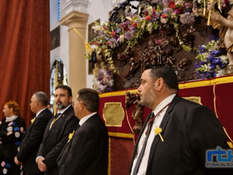 Un grupo de personas en trajes formales observan una procesión religiosa con una estatua en el centro, rodeada de flores y decoraciones elaboradas.