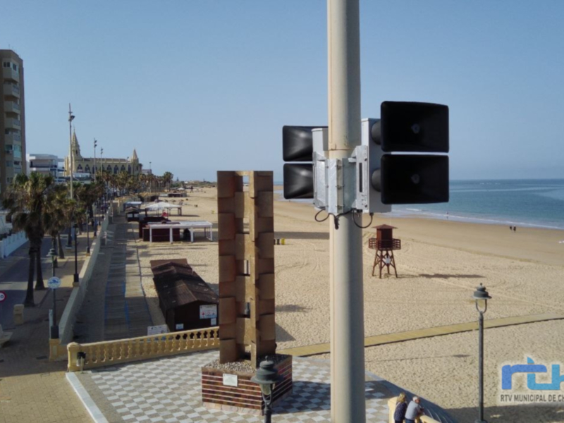 Imagen de una playa con edificios y una señal de tráfico en primer plano. La playa está desierta, con personas paseando y disfrutando del clima soleado. El horizonte se puede ver con el mar en la distancia.