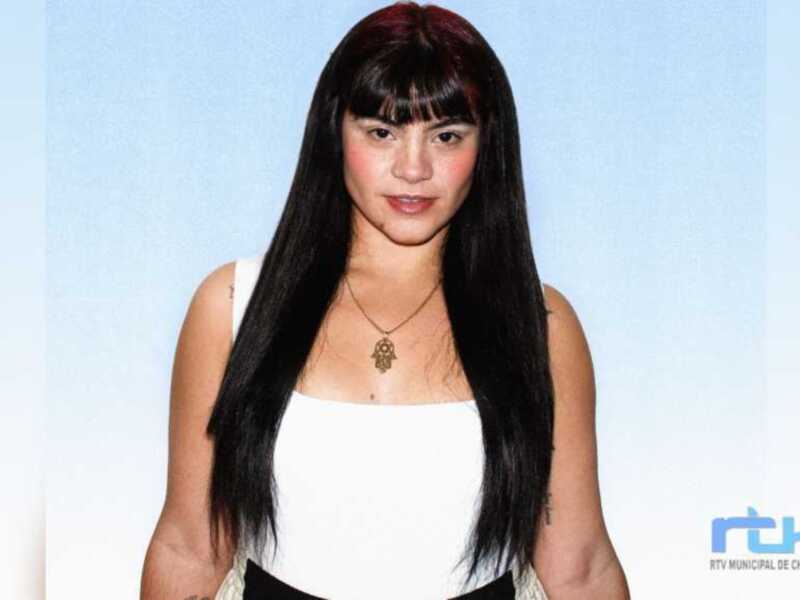 A una mujer con cabello largo y negro, peinado recto. Lleva un top blanco y una cadena con un símbolo en la garganta. La imagen es de fondo azul claro, y la marca "RTV MUNICIPAL DE CHIPONA" está en el fondo.