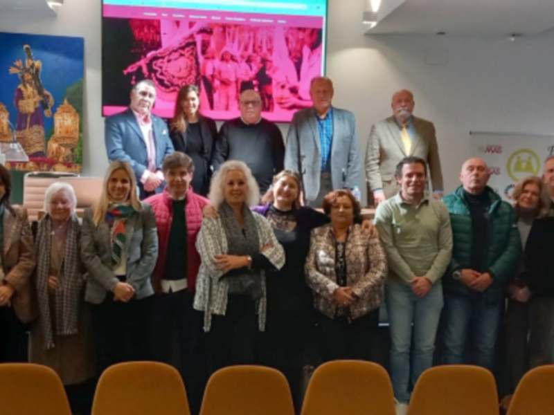 Una reunión formal en un salón con una pantalla de fondo que muestra información. La imagen incluye a varias personas, algunos con insignias y otros con carteles de apoyo. La decoración incluye una bandera y un cartel en el fondo, junto con sillas amarillas.
