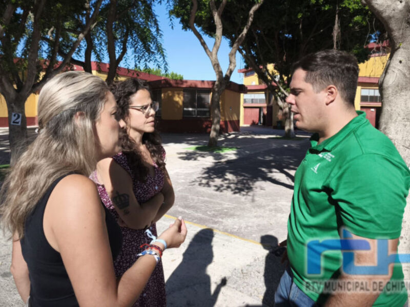 Tres personas conversan en un área de aparcamiento, con un edificio y árboles al fondo. La imagen muestra una escena casual en un entorno urbano.