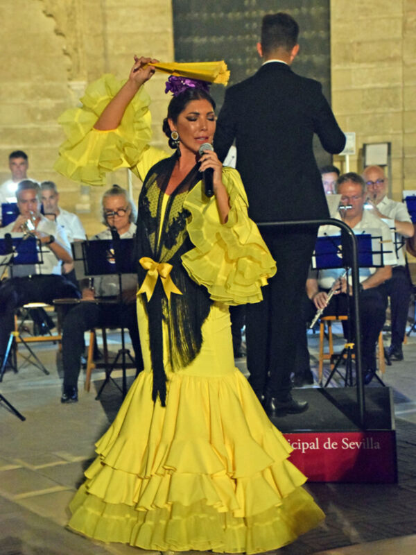 Una mujer en un traje de flamenca amarillo y negro, con un velo azul en la cabeza, canta frente a una orquesta. El escenario es al aire libre con un muro de piedra en el fondo y una placa roja que dice "Municipal de Sevilla".