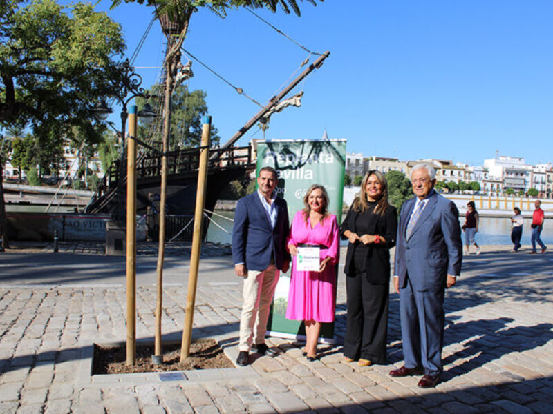 Una imagen de una familia posando frente a un monumento en un parque. La madre y el padre están sosteniendo una placa, mientras que los hijos están detrás de ellos. El parque tiene un camino adoquinado y árboles, con edificios en el fondo.