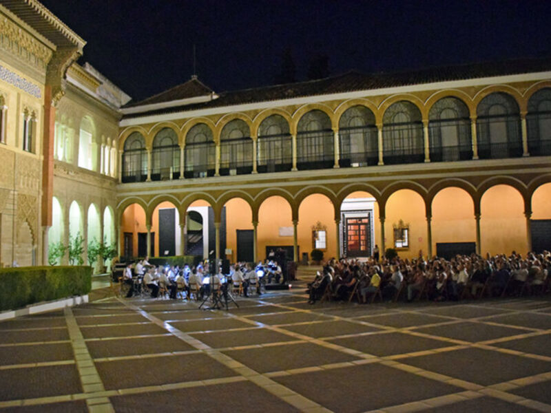 Un patio de noche con una arquitectura histórica, gente sentada en el suelo y un grupo de músicos tocando al fondo.