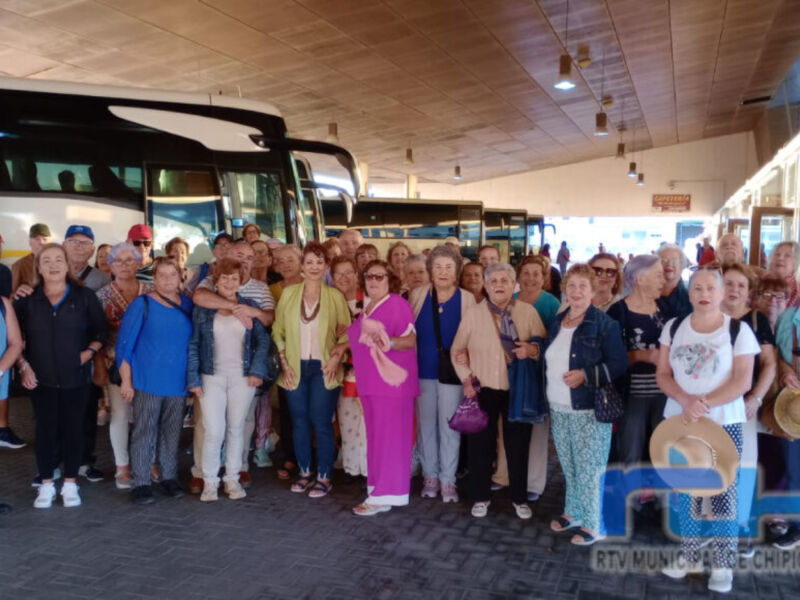 Una multitud de personas mayores se reúnen en un terminal de autobuses, posando para una foto colectiva. La imagen muestra a varias personas vestidas con ropa casual, algunos con sombreros y otros con bastones de caminata. El fondo incluye un autobús blanco y negro, iluminaciones en el techo y una puerta de cristal.