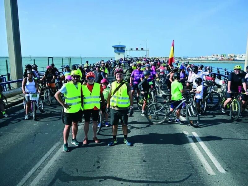 Una multitud de ciclistas con cascos y chalecos reflectantes se preparan para una carrera en un puente. La gente está dispuesta, algunos con bicicletas y otros con mochilas. El cielo es claro y el mar está visible en el fondo, creando una escena vibrante de deporte al aire libre.