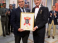 Dos hombres en trajes sostienen un diploma con un escudo heráldico. El fondo muestra otros individuos en trajes y una pared con retratos de personas.