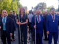 Una imagen de tres personas vestidas formalmente, posiblemente en un desfile o ceremonia oficial. Las personas llevan insignias y estandartes, lo que sugiere un contexto ceremonial o religioso. La escena parece ser al aire libre con árboles y cielo visible en el fondo.