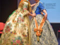 Dos mujeres en trajes tradicionales valencianos durante una actuación en el escenario de la Casa Regional Valenciana en Sevilla, con el logo de la Generalitat Valenciana.