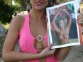 Una mujer con una camiseta rosa y pantalones negros sostiene un libro abierto que muestra a una mujer desnuda. La escena se desarrolla en un parque con árboles y una calle visible al fondo.