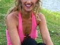 Una mujer con cabello rizado y largo, vistiendo una camiseta rosa sin mangas y pantalones negros, sentada en el césped frente a un lago.