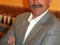 Un hombre con barba y bigote, vistiendo una chaqueta de lana y camisa blanca, con los brazos cruzados. Fondo amarillo con una manta a cuadros en la parte inferior.