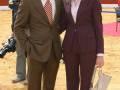 Dos personas posan en un estadio, uno con traje marrón y corbata roja, la otra con traje de chaqueta y pantalones a rayas. La imagen muestra una escena formal en un lugar público con gradas y un área de juego al fondo.