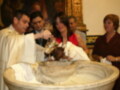 Un sacerdote bendice a un bebé recién nacido durante un bautismo, rodeado de familiares y amigos. La imagen muestra una ceremonia religiosa con un niño en el centro del bautismo, mientras el sacerdote realiza la bendición.