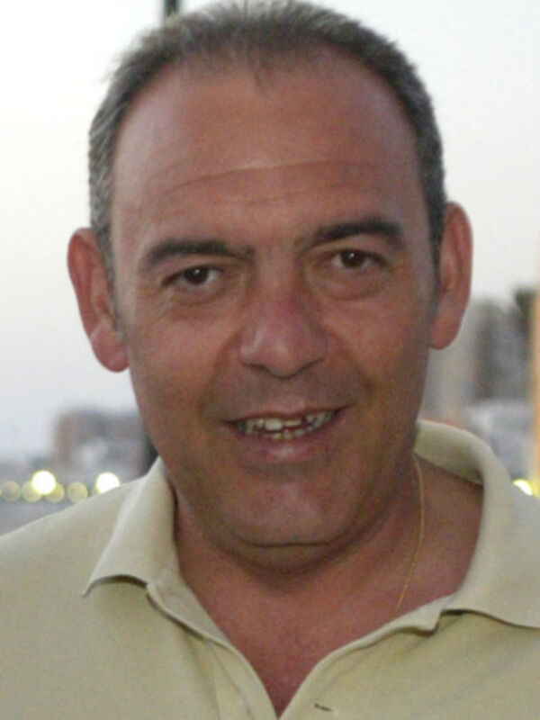 Un hombre con cabello corto y gris, sonriendo ampliamente. Está vestido con una camisa de color claro y lleva un collar largo. El fondo parece ser al aire libre con luces brillantes en el horizonte.