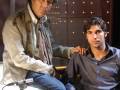 Dos hombres en un entorno de luces doradas, uno está sentado y el otro está ayudándolo con un objeto.