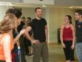 Un grupo de personas en una clase de danza, con un hombre en el centro sonriendo y dos mujeres a su lado riendo. La escena se desarrolla en un ambiente de gimnasia con espejos y una pista de baile visible.