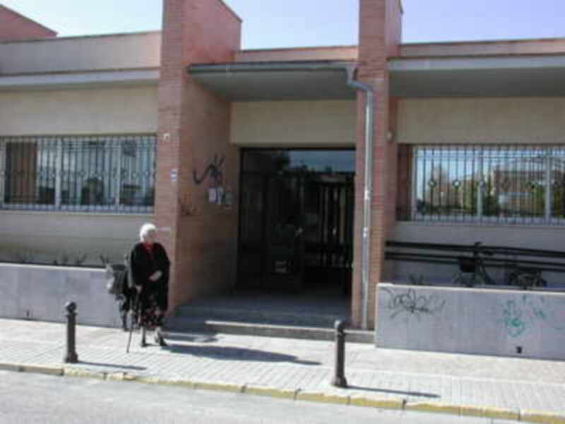 Una persona mayor camina con bastones frente a un edificio con ventanas y rejas, con grafiti en la pared.