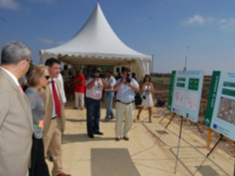 Una imagen de una reunión formal al aire libre con personas vestidas de negocios, un toldo blanco y carteles informativos en el fondo.