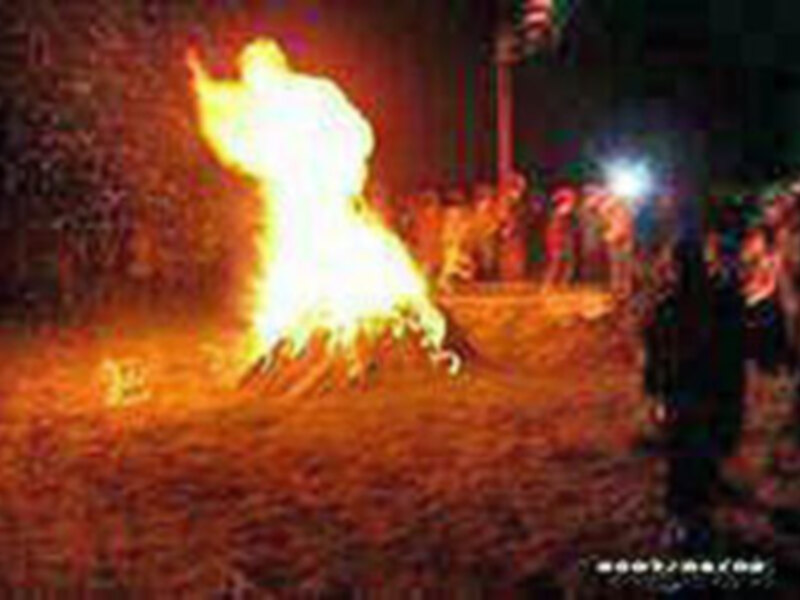 Una gran hoguera ardiendo en el centro de una noche oscura, rodeada por personas que observan la escena. La llama se eleva alto, iluminando el entorno y creando una atmósfera festiva.