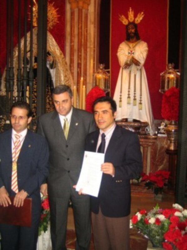 En una iglesia, tres hombres en trajes posan con un documento. La imagen incluye una estatua de Cristo y flores rojas en el fondo.
