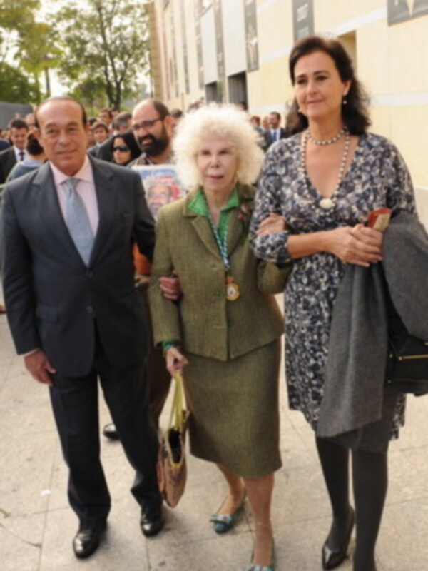 Una mujer mayor con un abrigo verde y una medalla en el cuello camina junto a un hombre con traje negro, seguida por una mujer con vestido floral y abrigo gris. Todos parecen estar en un evento formal, posiblemente una celebración o reunión importante.