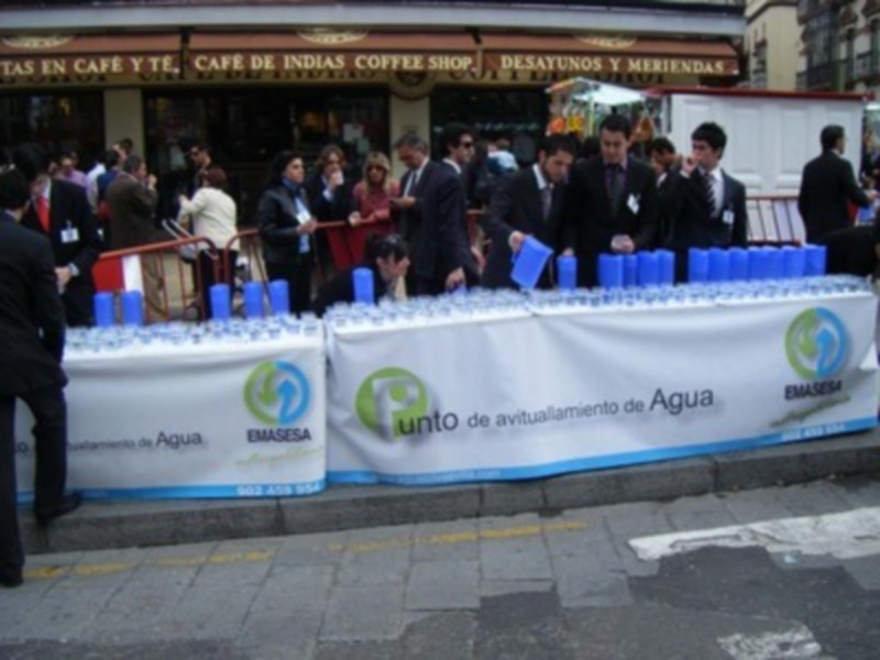 Un stand de agua embotellada en una calle, con personas en el fondo. El stand tiene una bandera blanca con el logo de EMASESA y el texto 'punto de reutilización de Agua'.