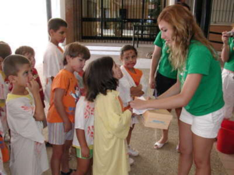 Niños esperando en línea para recibir una caja de comida. Adulto con cabello largo y verde distribuyendo las cajas a los niños.