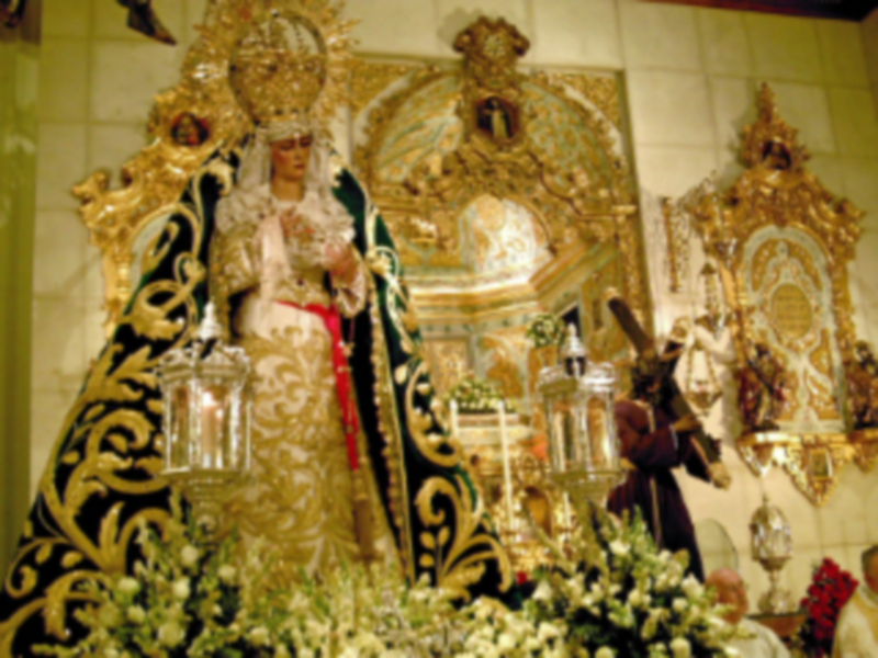 Imagen religiosa con una Virgen en el centro, rodeada de ornamentos dorados y flores blancas. En la parte superior se observa un altar con estatuas y adornos, todo ello en un ambiente de iglesia.