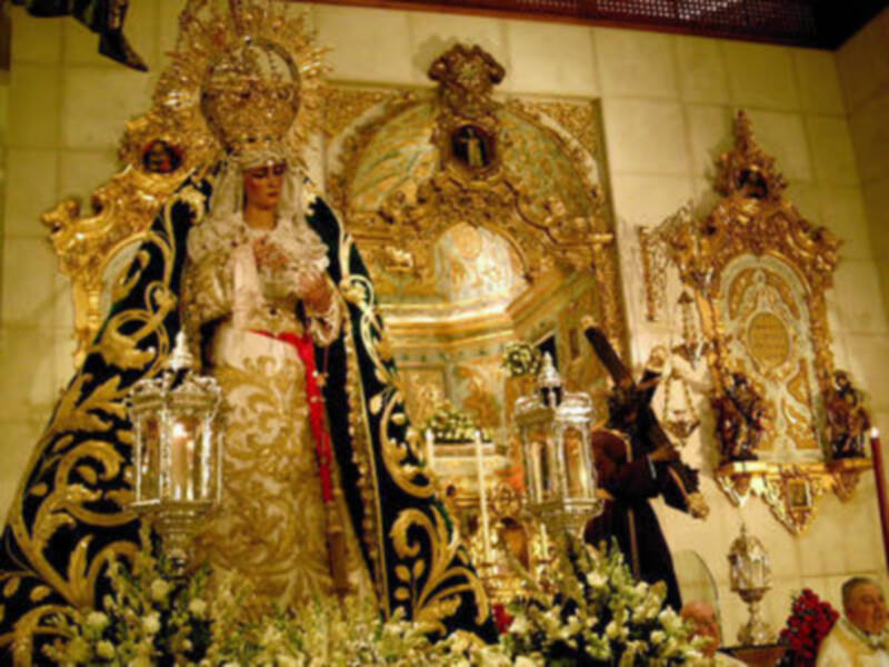 Imagen de una Virgen María vestida con ornamentos y vestidos, rodeada por flores y objetos religiosos en un altar.