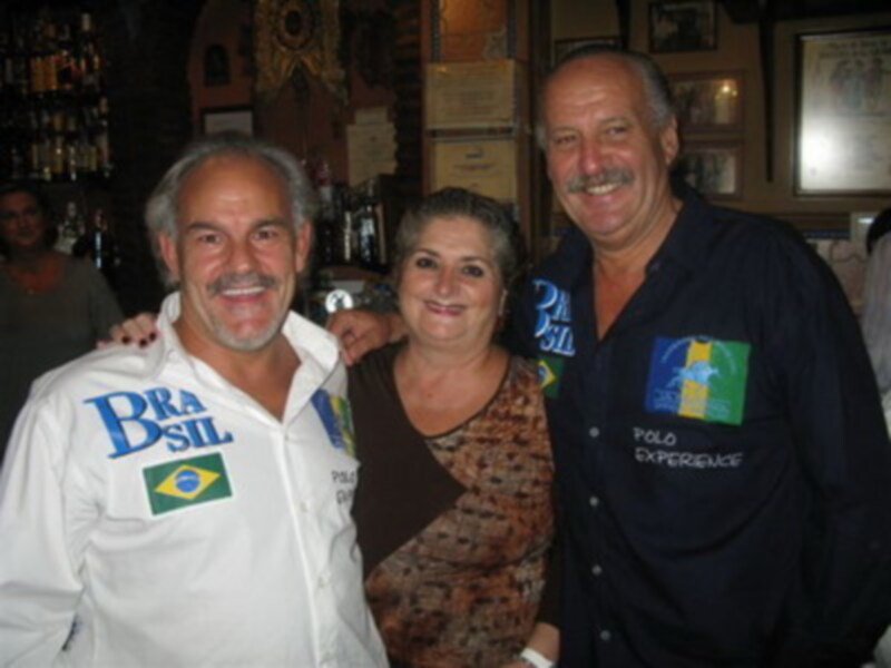 Tres personas sonrientes, dos hombres y una mujer, posando en un ambiente que parece ser un bar o pub. Los hombres llevan camisetas con el logo de Brasil, mientras que la mujer está en medio. La decoración incluye barras de bebidas y carteles en el fondo.