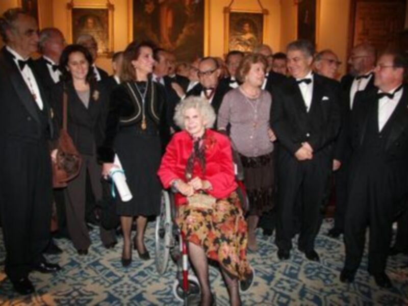 Una anciana en silla de ruedas, vestida con un abrigo rojo y falda floral, sonríe mientras se encuentra entre una multitud de personas vestidas con trajes formales. La escena parece ser un evento formal, posiblemente una gala o recepción, con una alfombra de color verde y un fondo que incluye cuadros y decoración.